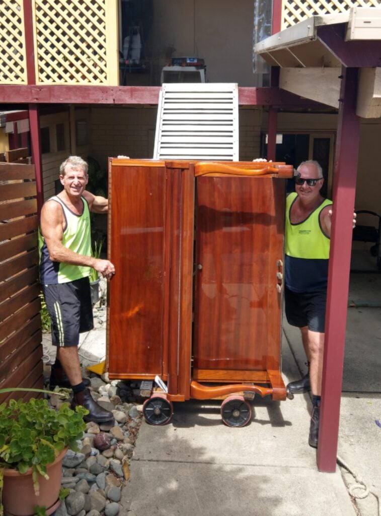 Two men of Countrycare Movers in high-visibility tank tops standing outdoors on a patio, holding a large, polished furniture that is being moved on a wheeled dolly.