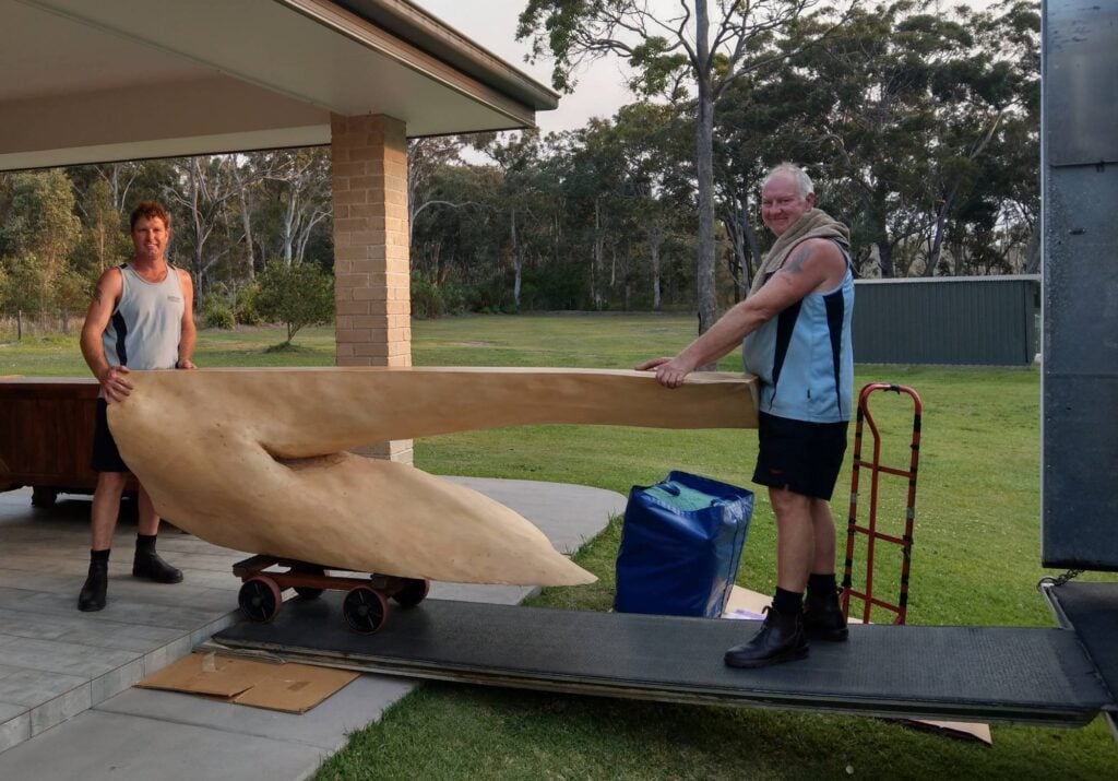 Two movers in blue uniforms carefully guide a large, uniquely shaped wooden sculpture down a truck ramp using a small dolly.