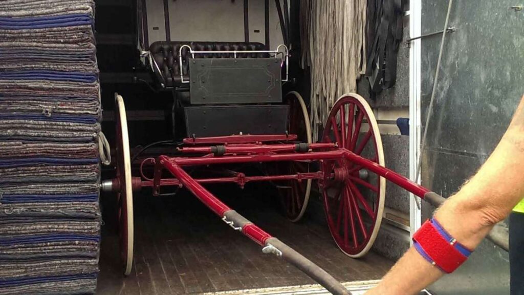 A red horse-drawn carriage being loaded into or unloaded from a transport truck, positioned next to a stack of moving blankets.